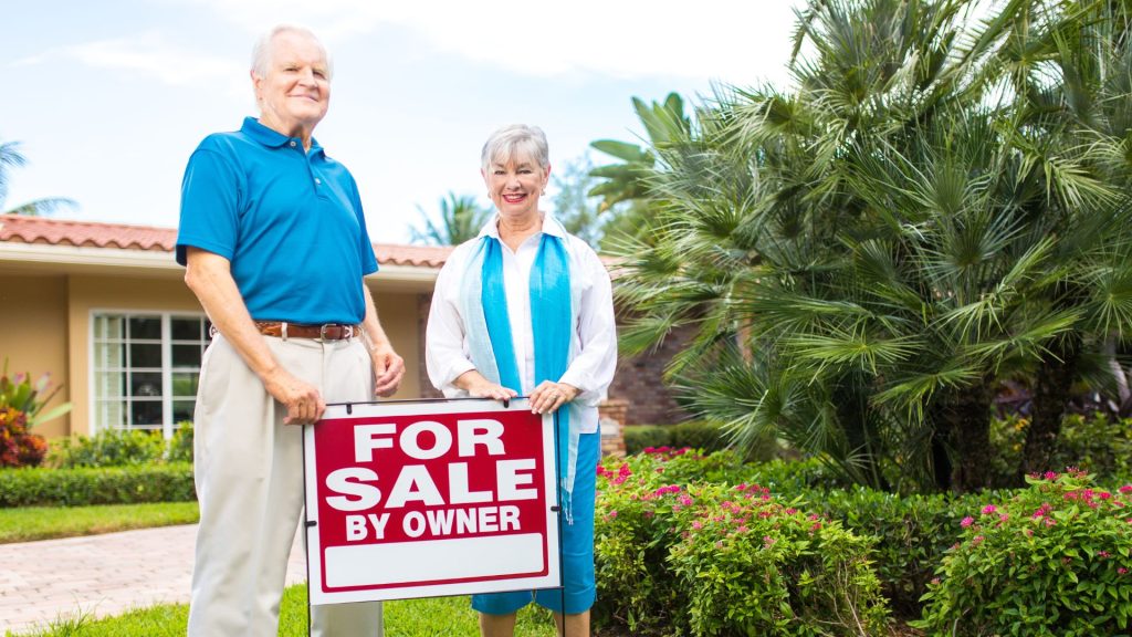 An elderly couple in front of a house, holding down a signboard that says "For Sale"