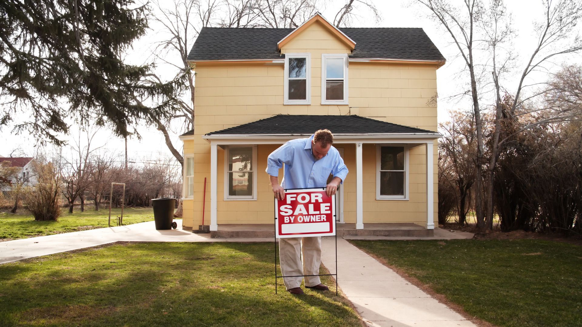 A man holding down a "For Sale" signboard with his house behind him