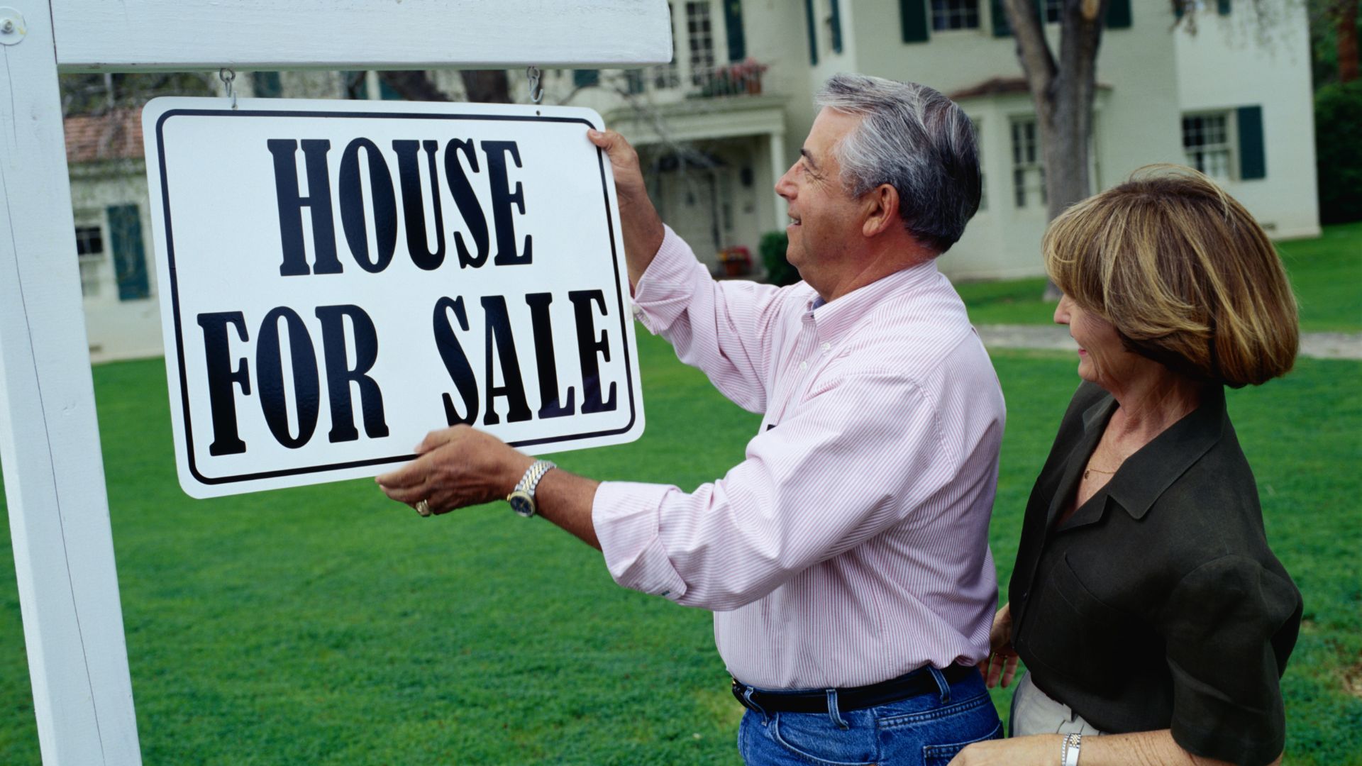 An elderly couple putting up a "House for sale" sign in front of their house